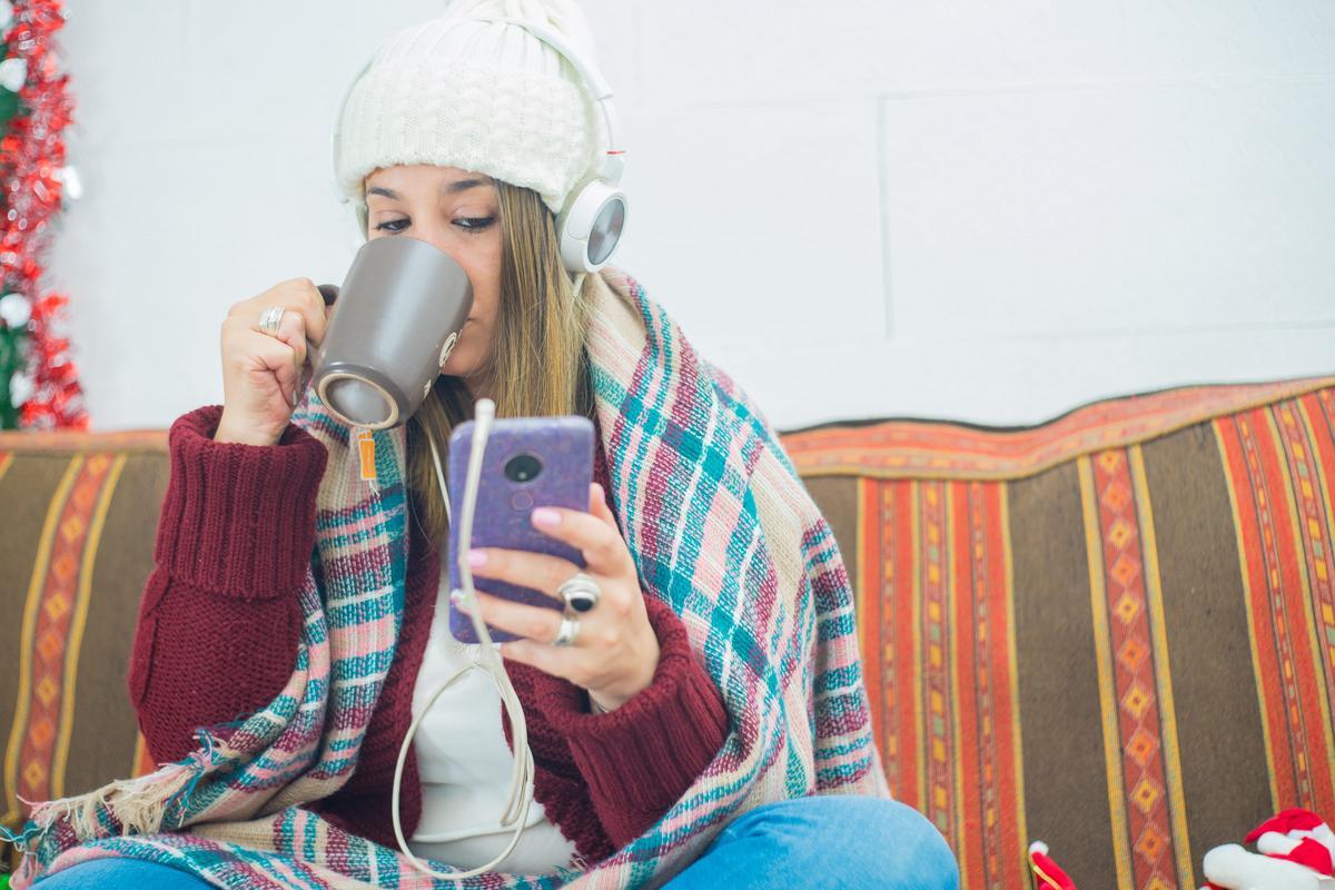 Woman wrapped in a blanket and winter hat checking her phone at home